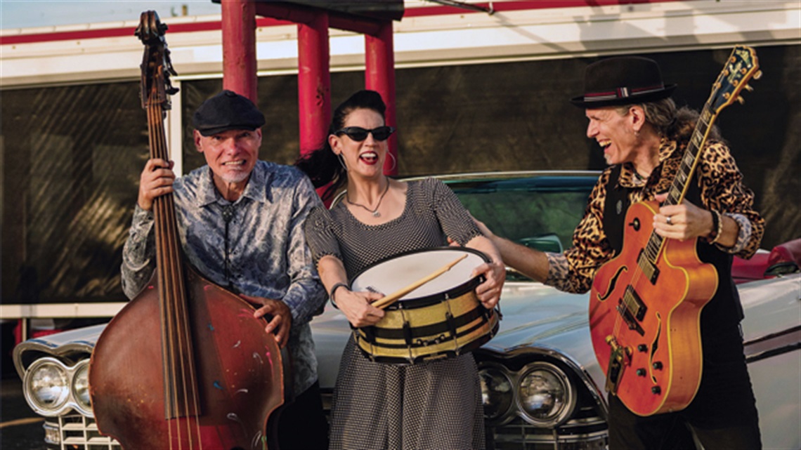 Photo of 3-member band The Mighty Flea Circus holding instruments, laughing, in front of a vintage car.