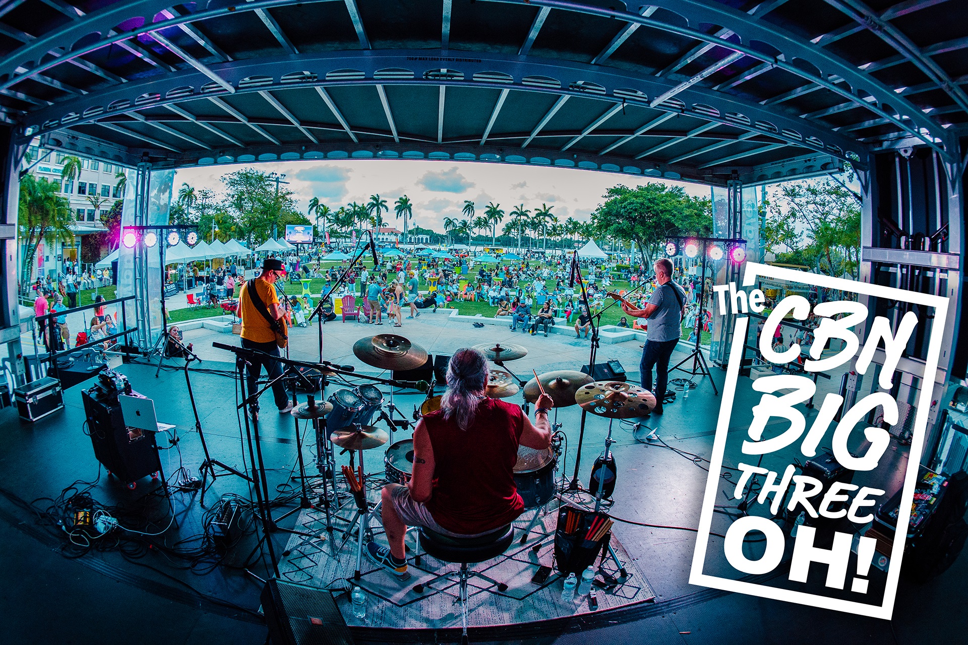 Photo of concert taken from the back of the stage looking out onto the lawn. Musicians are in foreground and crowd beyond the stage in the distance. The CBN Big Threek-OH! stamp is in the bottom right corner.