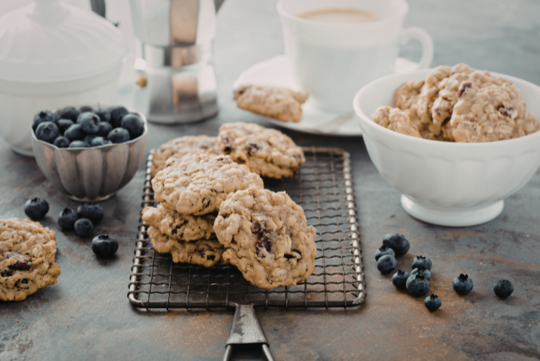 Blueberry oatmeal cookies cooling on a wire rack, surrounded by fresh blueberries, with additional cookies in a white bowl and a cup of coffee in the background.