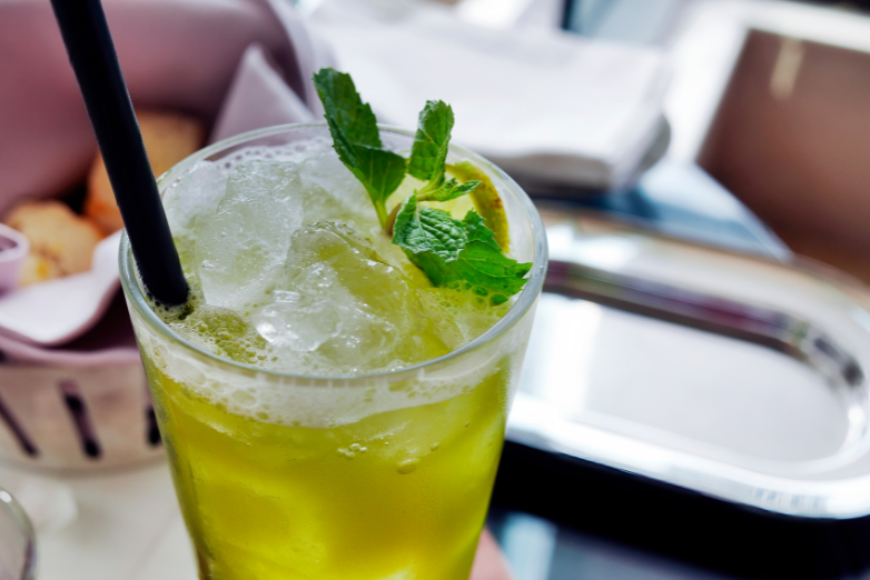 A cold yellow green beverage served in a tall glass packed with ice cubes and garnished with fresh green mint leaves and lime slices.