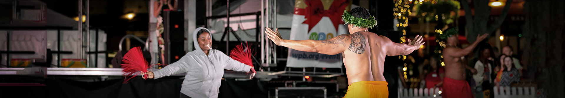 Pano photo of a laughing patron echoing the movements of a Polynesian dancer, arms outstretched, holding hand tassels.